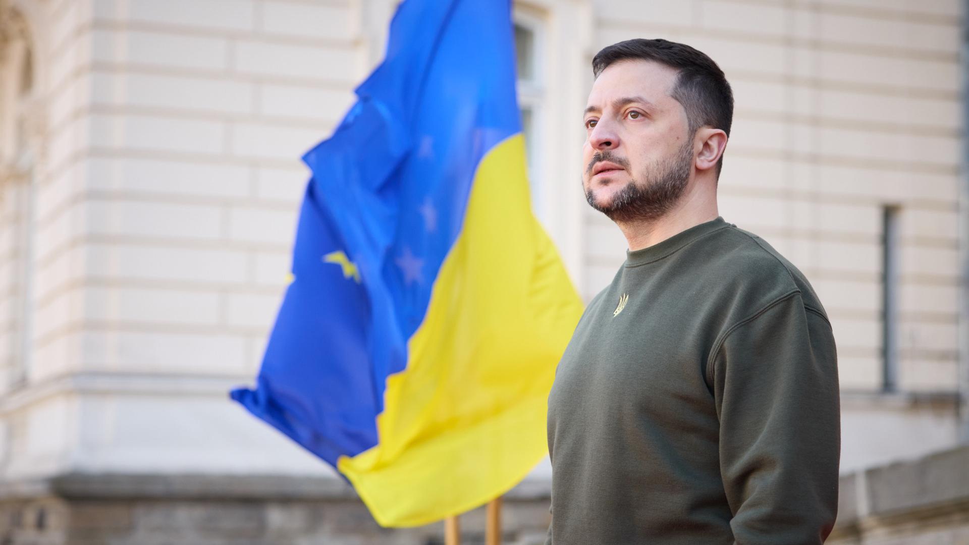 Der ukrainische Präsident Wolodymyr Selenskyj steht vor einem Gebäude, neben ihm weht eine ukrainische Flagge