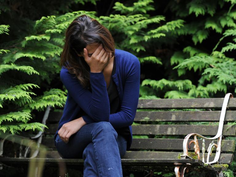 Eine junge Frau sitzt auf einer Bank und hält eine Hand vor ihr Gesicht. 
