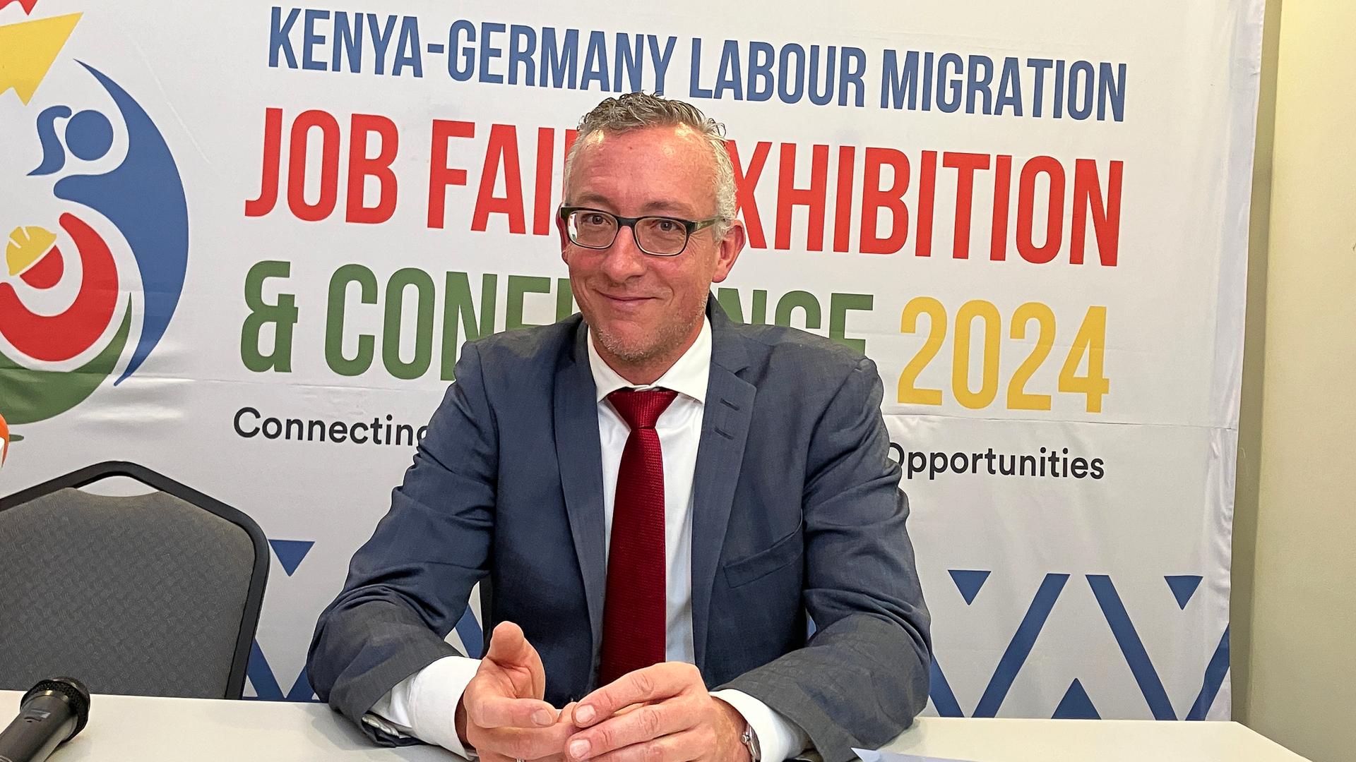 Sebastian Groth, deutscher Botschafter in Kenia, sitzt an einem Tisch. Hinter ihm ist ein Plakat, auf dem oben in blauer Schrift "Kenya-Germany Labour Migration" steht. 