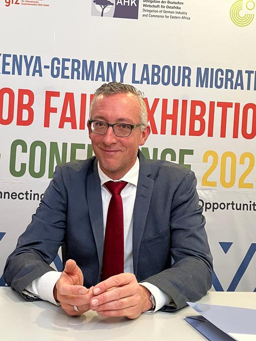 Sebastian Groth, deutscher Botschafter in Kenia, sitzt an einem Tisch. Hinter ihm ist ein Plakat, auf dem oben in blauer Schrift "Kenya-Germany Labour Migration" steht. 