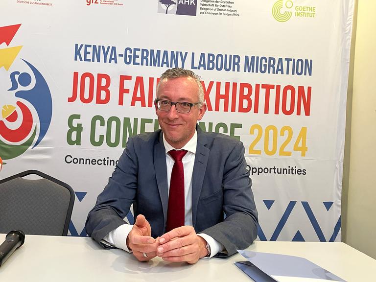 Sebastian Groth, deutscher Botschafter in Kenia, sitzt an einem Tisch. Hinter ihm ist ein Plakat, auf dem oben in blauer Schrift "Kenya-Germany Labour Migration" steht. 