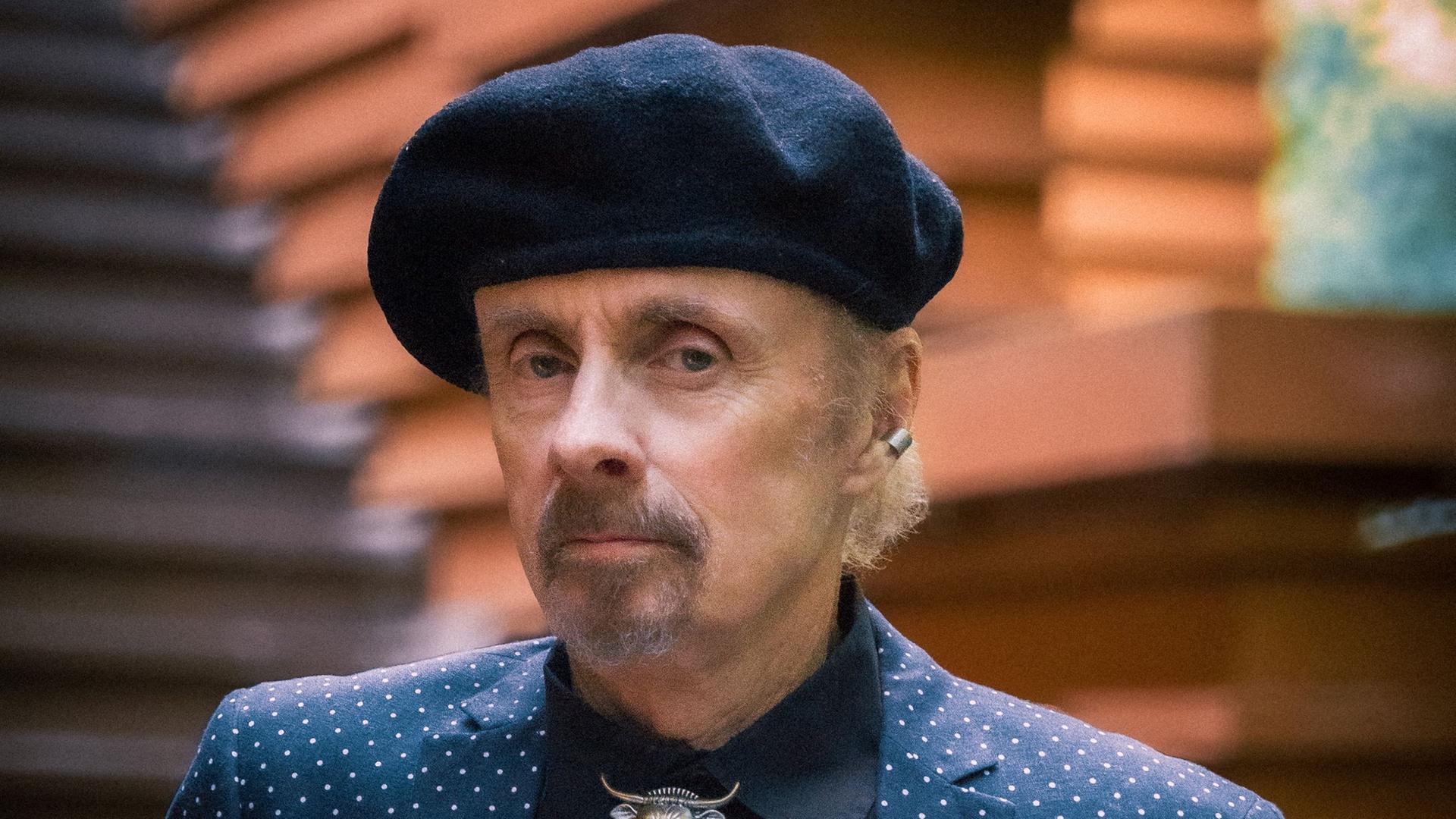 Der Autor T.C. Boyle schaut ernst in die Kamera. Er hat einen kurzen, grau melierten Bart und trägt eine schwarze Baskenmütze, ein schwarzes Hemd und darüber ein blaues Jackett mit weißen Punkten.