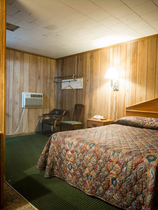 Ein Zimmer in einem günstigen Motel. Mit falscher Holzvertäfelung und grünem Teppich unter dem geblümten Bett.