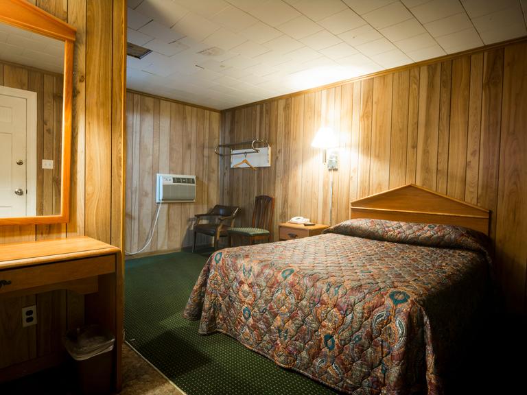 Ein Zimmer in einem günstigen Motel. Mit falscher Holzvertäfelung und grünem Teppich unter dem geblümten Bett.