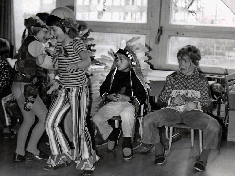Verkleidete weiße Kinder feiern Fasching in einem Klassenzimmer. Zwei Kinder tanzen, ein schwarzer Junge im Indianerkostüm sitzt traurig auf einem Stuhl daneben. 