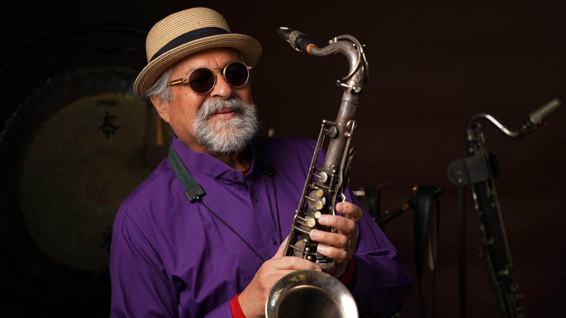 Ein Mann mit Strohhut und Sonnenbrille im lilafarbenen Hemd zeigt sein silbernes Saxofon in die Kamera.