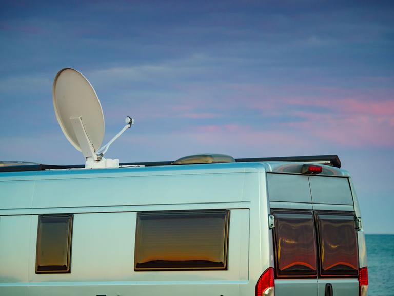 Ein Camper mit Satellitenschüssel auf dem Dach vor einem Sonnenuntergang am Meer.