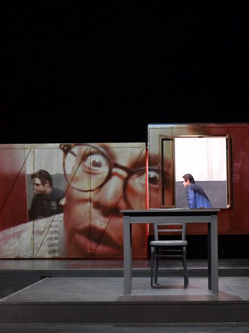 Bild der Bühne einer Aufführung von "Jugend ohne Gott" am Volkstheater Rostock. Im Hintergrund des Bühnenraums stehen Stellwände, die von einem übergroßen Gesicht bedeckt sind. Im Vordergrund Tische, am rechten Rand sitzt eine Frau im roten Kostüm auf einem Tisch und begutachtet die Szene. Am linken Bühnenrand steht ein Mann. 