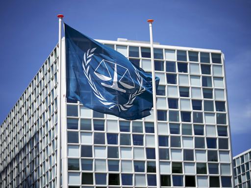 Außenansicht des Internationalen Strafgerichtshofs in Den Haag. Davor eine blaue Flagge, die in weiß das Logo des Gerichts abbildet: eine Waage, die von einem Lorbeerkranz umgeben ist.