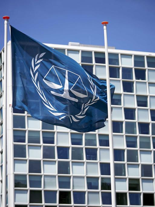 Außenansicht des Internationalen Strafgerichtshofs in Den Haag. Davor eine blaue Flagge, die in weiß das Logo des Gerichts abbildet: eine Waage, die von einem Lorbeerkranz umgeben ist.