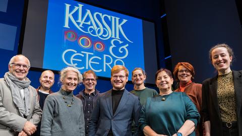 Neun Personen stehen auf der Bühne im Deutschlandfunk Kammermusiksaal in Köln und blicken lächelnd in die Kamera. Im Hintergrund ist ein Visual mit dem Schriftzug "Klassik-Pop-et cetera" auf einer Leinwand zu sehen.