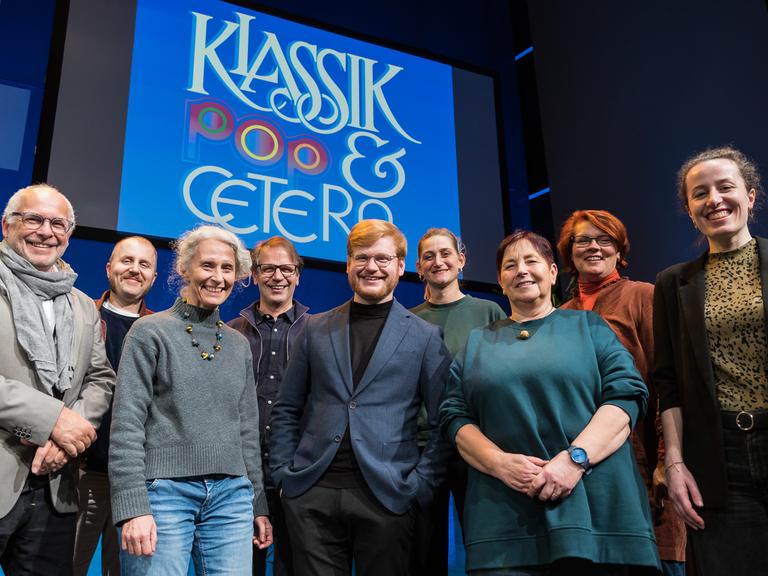 Neun Personen stehen auf der Bühne im Deutschlandfunk Kammermusiksaal in Köln und blicken lächelnd in die Kamera. Im Hintergrund ist ein Visual mit dem Schriftzug "Klassik-Pop-et cetera" auf einer Leinwand zu sehen.