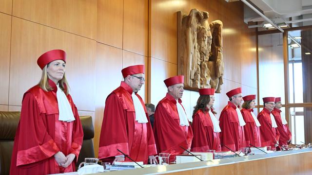 Richterinnen und Richter in roter Kleidung und mit roter Kappe stehen in einer Reihe hinter einem Tisch. Hinter ihnen an der Wand ist eine Skulptur des Bundesadlers.