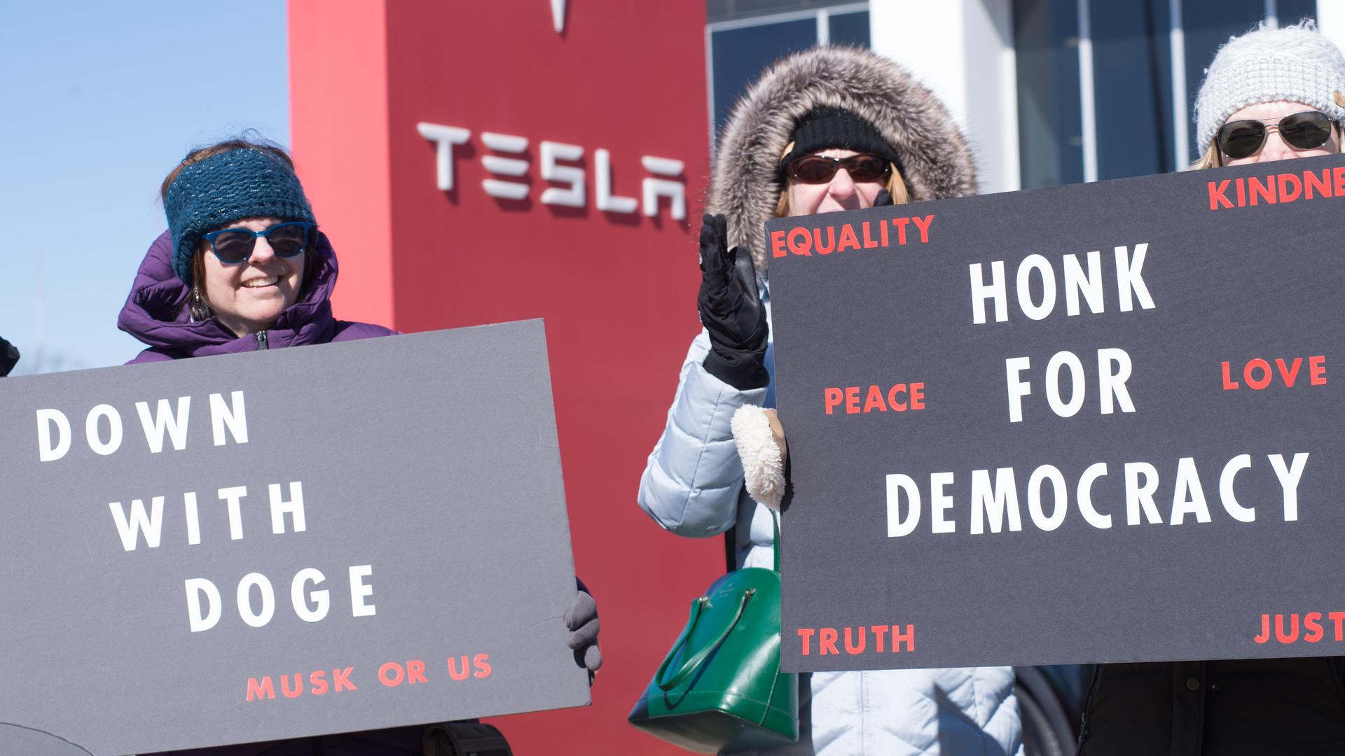 Drei Frauen mit Sonnenbrillen stehen vor der Tesla-Filiale und halten Schilder, die sich gegen "Doge" richten. 