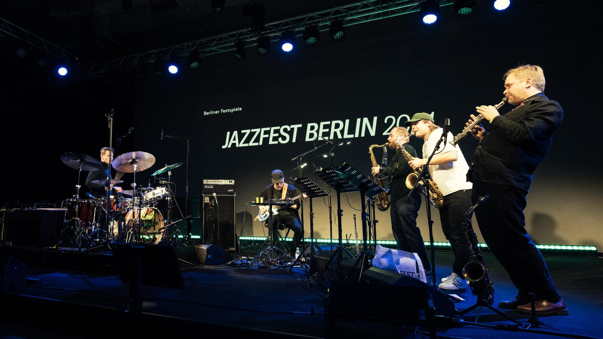 Eine Band aus, von links, Schlagzeuger, E-Bassist und drei Saxofonisten spielt auf einer Bühne. Im Hintergrund der Schriftzug "Jazzfest Berlin".