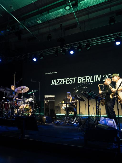 Eine Band aus, von links, Schlagzeuger, E-Bassist und drei Saxofonisten spielt auf einer Bühne. Im Hintergrund der Schriftzug "Jazzfest Berlin".