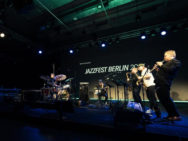 Eine Band aus, von links, Schlagzeuger, E-Bassist und drei Saxofonisten spielt auf einer Bühne. Im Hintergrund der Schriftzug "Jazzfest Berlin".