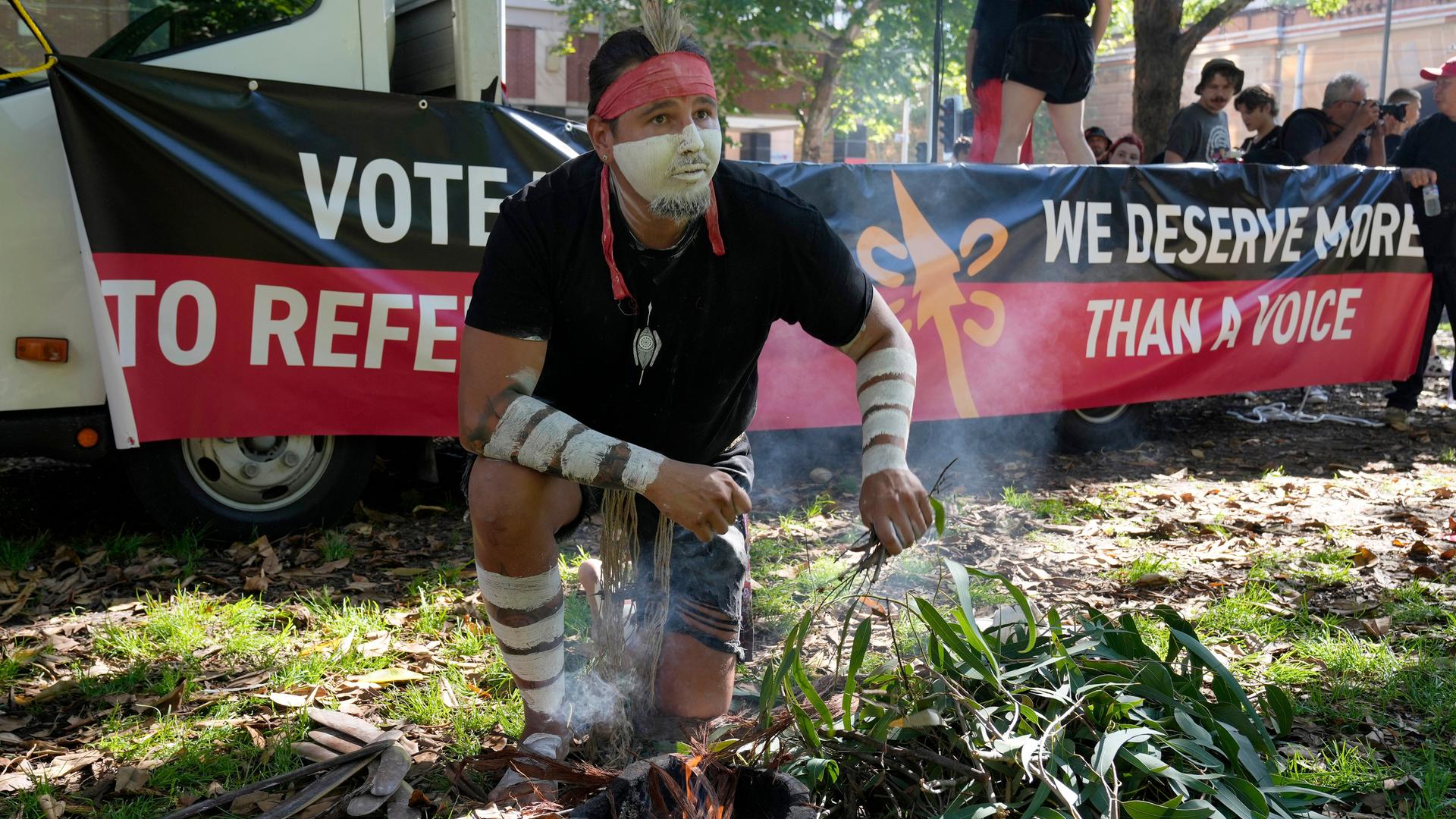 Ein Aborigine mit weißer Gesichtsbemalung entfacht ein zeremonielles Feuer. Hinter ihm steht auf einem Spruchband: "We deserve more than a voice." 