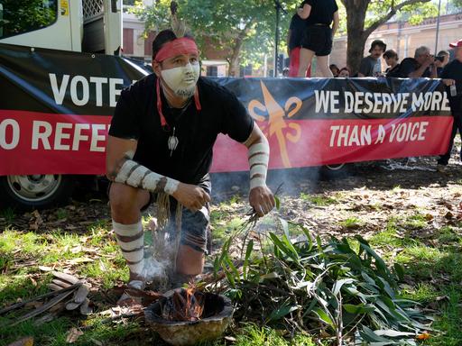 Ein Aborigine mit weißer Gesichtsbemalung entfacht ein zeremonielles Feuer. Hinter ihm steht auf einem Spruchband: "We deserve more than a voice." 