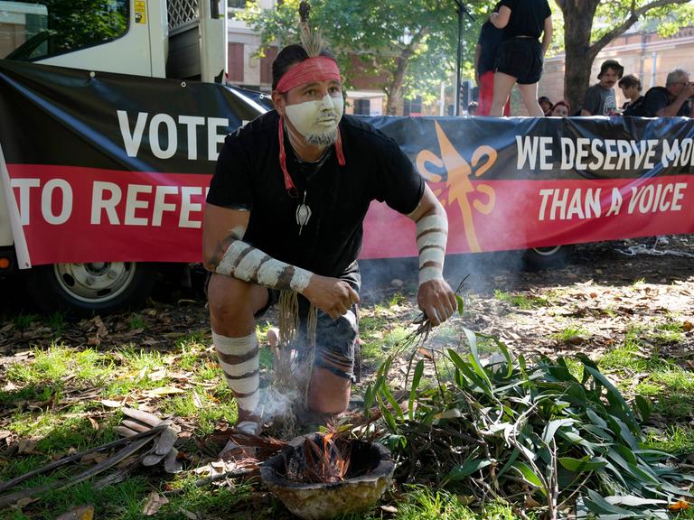 Ein Aborigine mit weißer Gesichtsbemalung entfacht ein zeremonielles Feuer. Hinter ihm steht auf einem Spruchband: "We deserve more than a voice." 
