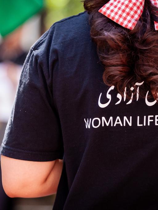 Das Foto zeigt eine Frau von hinten. Auf ihrem T-Shirt steht auf Englisch "Frau, Leben, Freiheit".