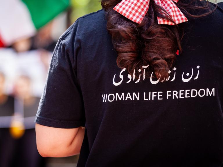 Das Foto zeigt eine Frau von hinten. Auf ihrem T-Shirt steht auf Englisch "Frau, Leben, Freiheit".