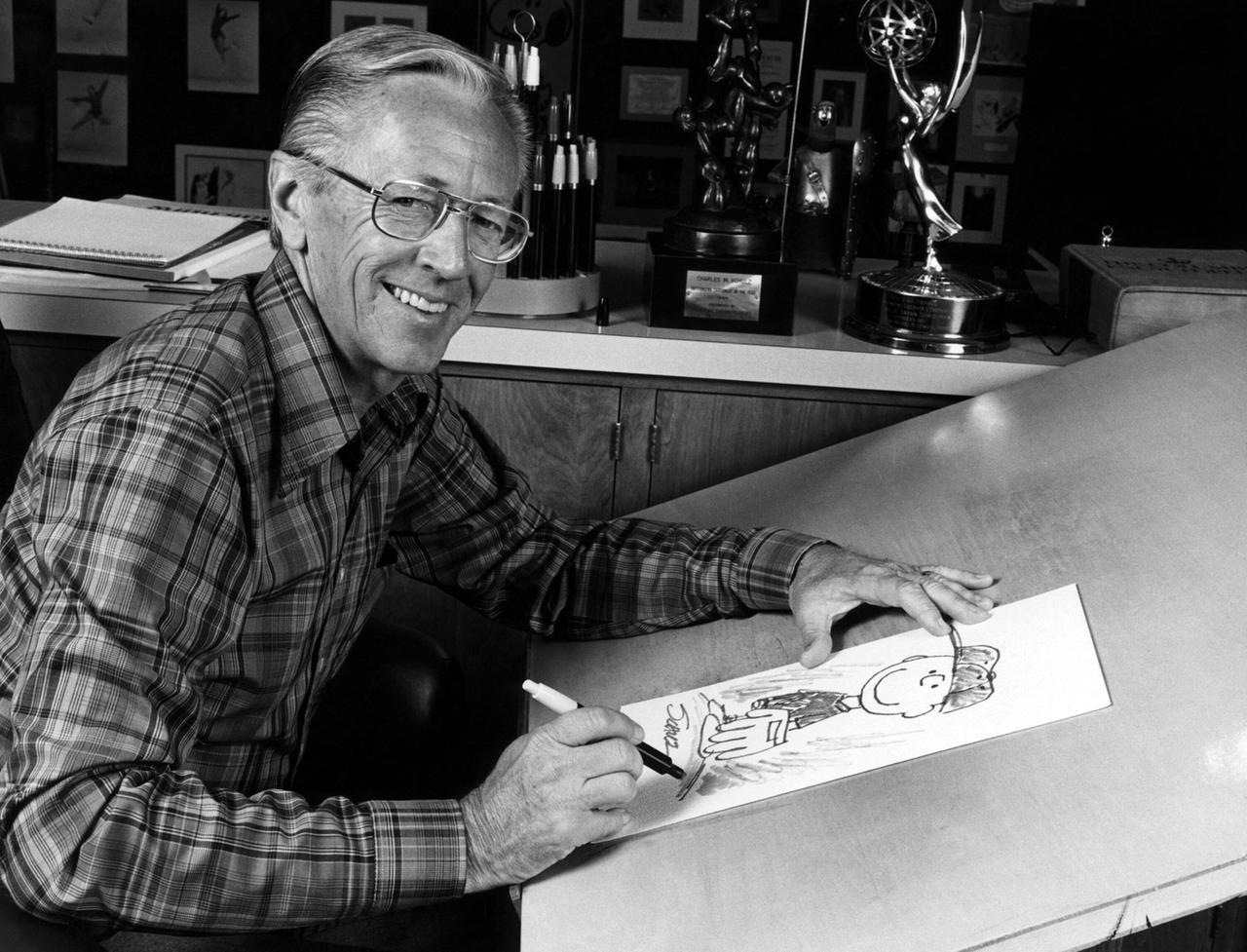 Charles M. Schulz sitzt lachend an seinem Zeichentisch, auf dem eine Skizze von Charlie Brown zu sehen ist