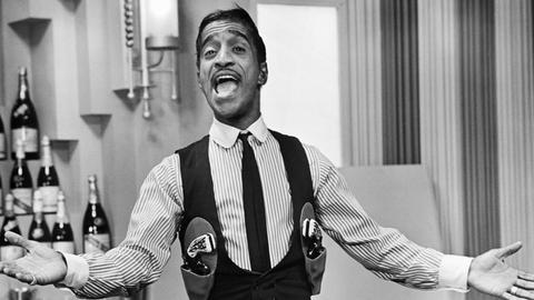 Schwarz-weiss Porträt von Sammy Davis Jr. mit Pistolengurt. Er singt und hat die Arme ausgebreitet.