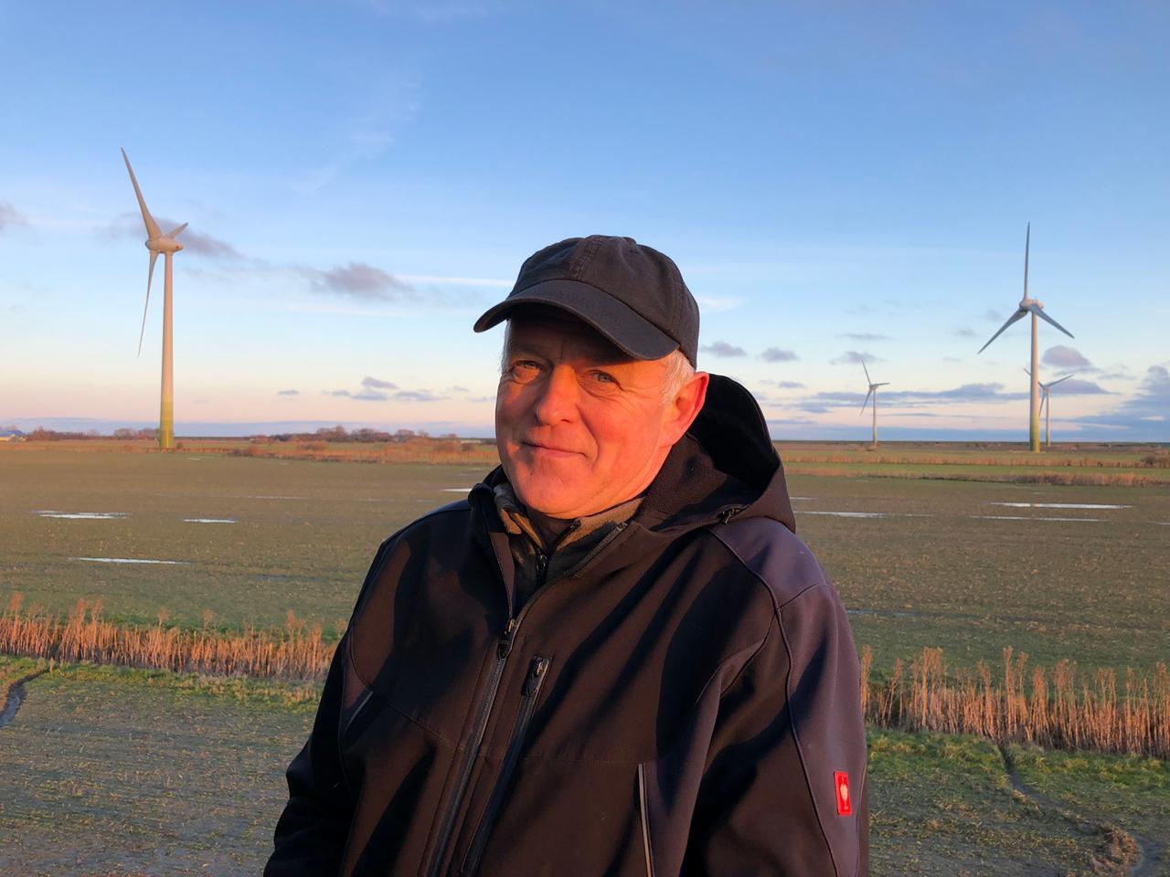 Kai Edlefsen steht mit Schirmmütze und dicker Jacke vor einem Feld, auf dem Windkraftanlagen stehen.
