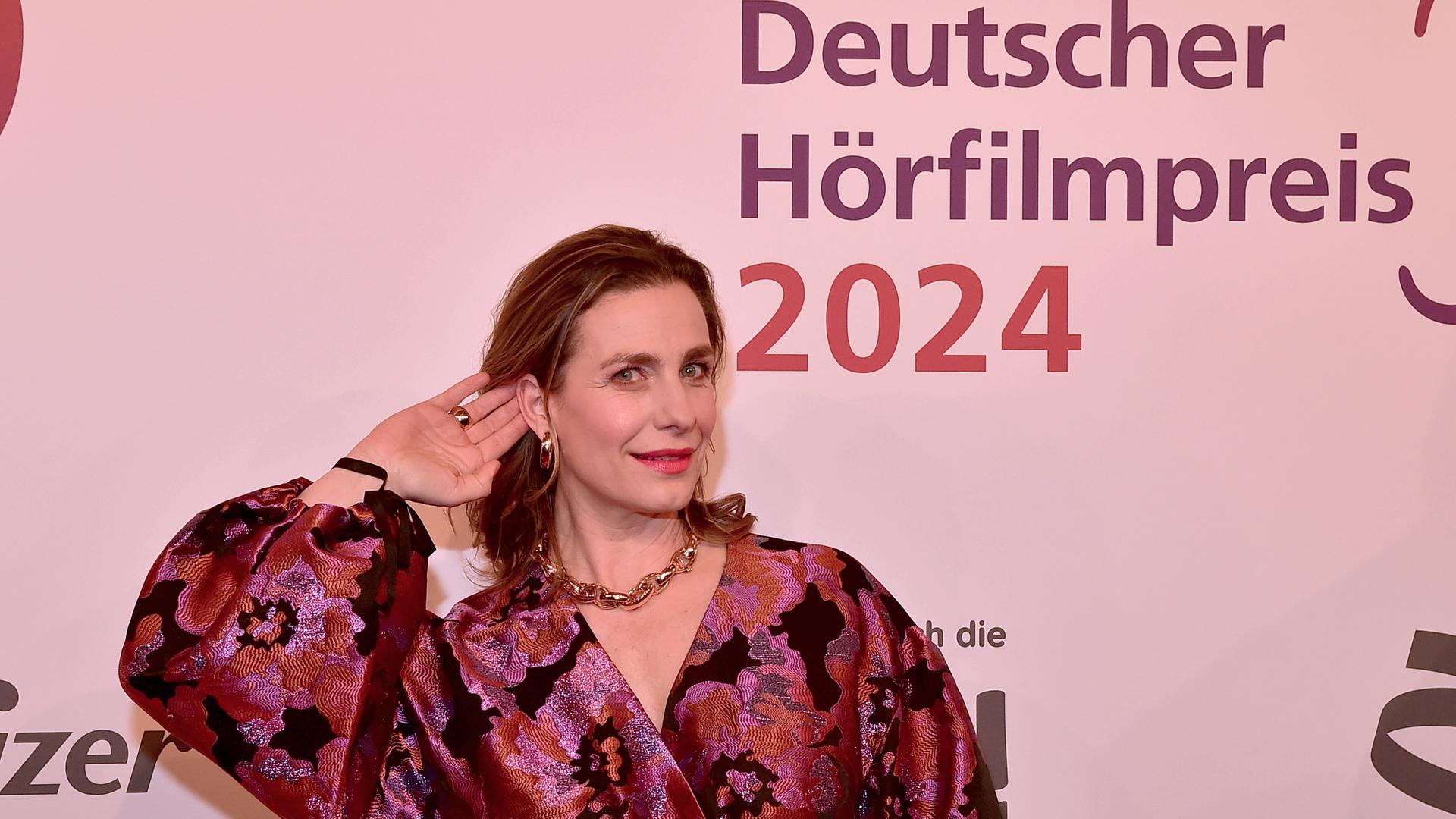 Die Schauspielerin Nadine Heidenreich steht beim 22. HörfilmPreis 2024 im Abendkleid auf dem roten Teppich und hält ihre Hand ans Ohr. 