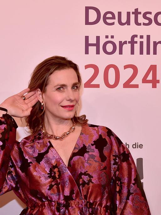 Die Schauspielerin Nadine Heidenreich steht beim 22. HörfilmPreis 2024 im Abendkleid auf dem roten Teppich und hält ihre Hand ans Ohr. 