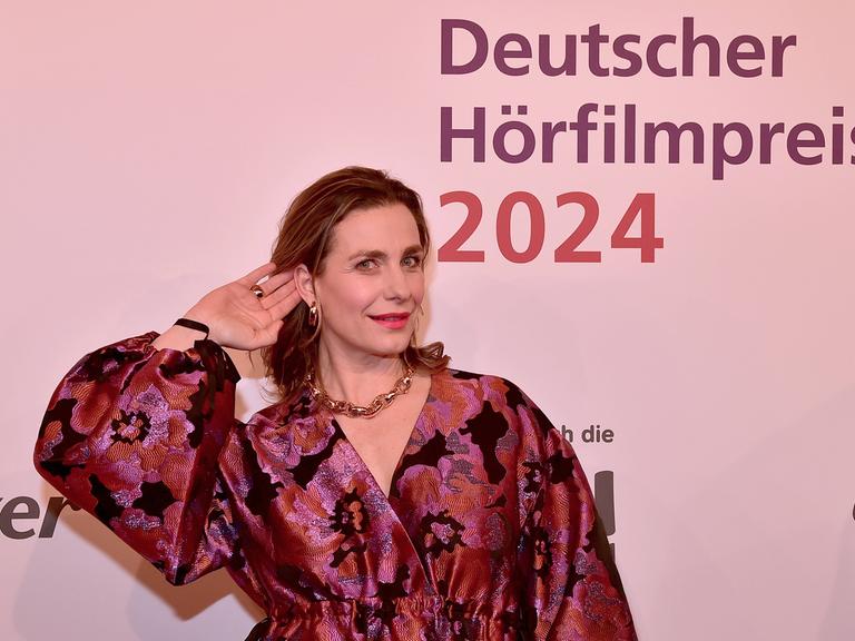 Die Schauspielerin Nadine Heidenreich steht beim 22. HörfilmPreis 2024 im Abendkleid auf dem roten Teppich und hält ihre Hand ans Ohr. 