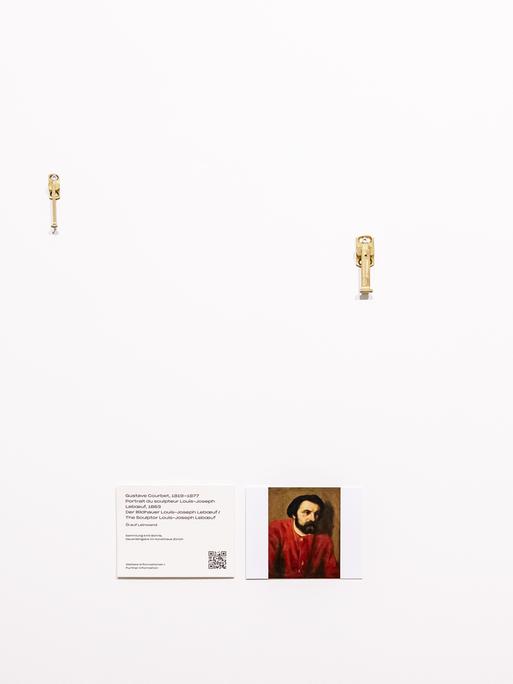 Eine leere Wand mit Aufäengungsvorrichtungen und einer kleinen Informationstafel am Platz wo das Bild "Portrait du Sculpteur Louis-Joseph" von Gustave Courbet abgehaengt wurde