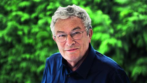 Ein Mann mit grauen Haaren und runder Brille steht im Grünen. Er trägt ein dunkelblaues Hemd und lächelt freundlich.