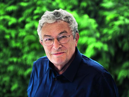 Ein Mann mit grauen Haaren und runder Brille steht im Grünen. Er trägt ein dunkelblaues Hemd und lächelt freundlich.