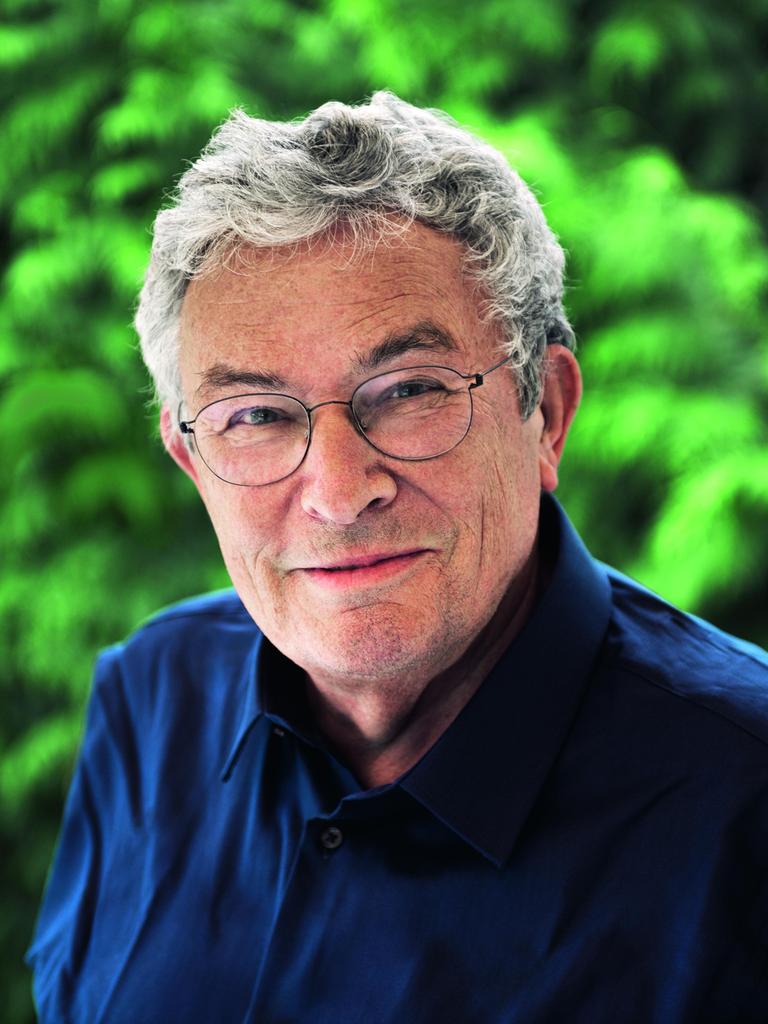 Ein Mann mit grauen Haaren und runder Brille steht im Grünen. Er trägt ein dunkelblaues Hemd und lächelt freundlich.