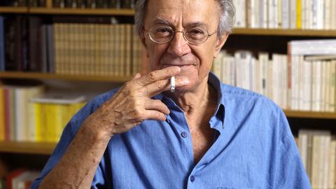 Jean-François Lyotard steht vor einem Bücherregal, raucht Zigarette und schaut lächelnd in die Kamera.