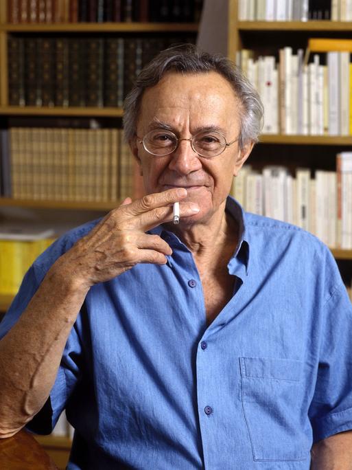 Jean-François Lyotard steht vor einem Bücherregal, raucht Zigarette und schaut lächelnd in die Kamera.