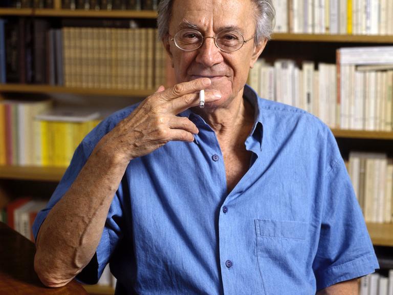 Jean-François Lyotard steht vor einem Bücherregal, raucht Zigarette und schaut lächelnd in die Kamera.