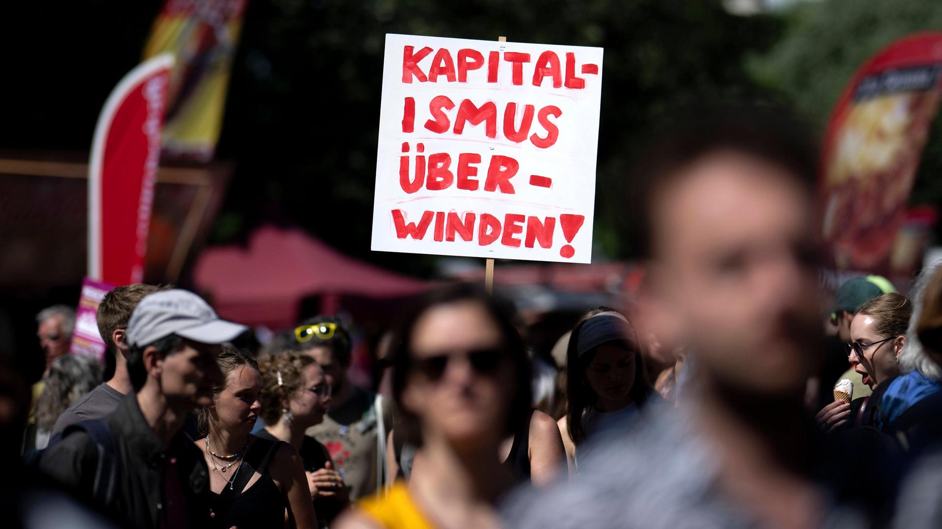 Das Bild zeigt viele Menschen, die bei einer Demonstration mitlaufen. In der Bildmitte ragt ein weißes Schild aus der Menge. Darauf steht in Rot: “Kapitalismus überwinden!” Das Bild zeigt viele Menschen, die bei einer Demonstration mitlaufen. In der Bildmitte ragt ein weißes Schild aus der Menge. Darauf steht in Rot: “Kapitalismus überwinden!”