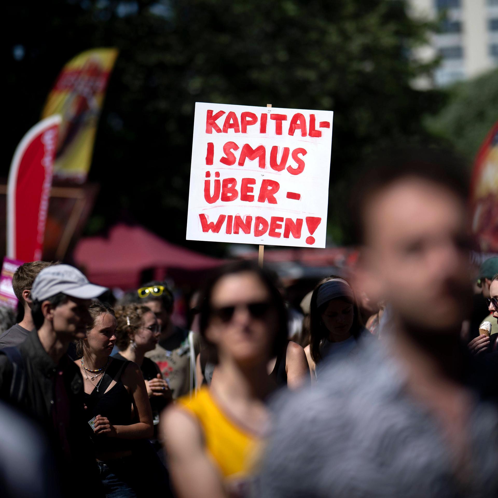 Das Bild zeigt viele Menschen, die bei einer Demonstration mitlaufen. In der Bildmitte ragt ein weißes Schild aus der Menge. Darauf steht in Rot: “Kapitalismus überwinden!” 