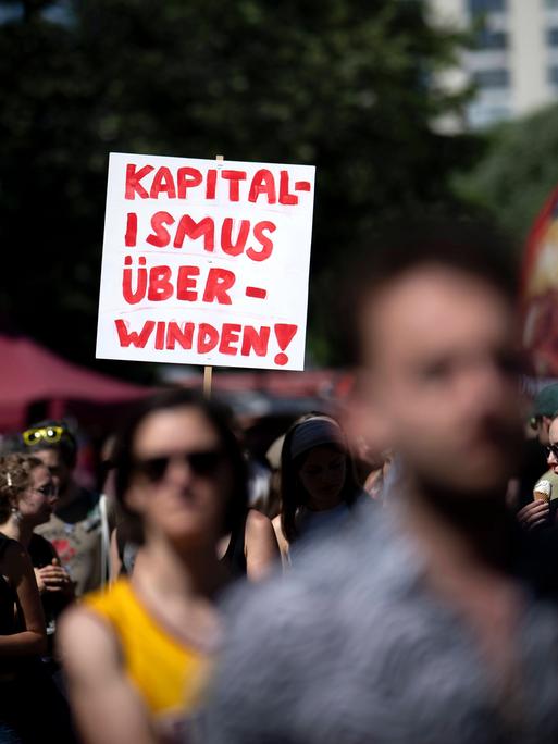 Das Bild zeigt viele Menschen, die bei einer Demonstration mitlaufen. In der Bildmitte ragt ein weißes Schild aus der Menge. Darauf steht in Rot: “Kapitalismus überwinden!” 