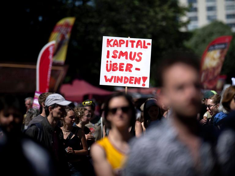 Das Bild zeigt viele Menschen, die bei einer Demonstration mitlaufen. In der Bildmitte ragt ein weißes Schild aus der Menge. Darauf steht in Rot: “Kapitalismus überwinden!” 