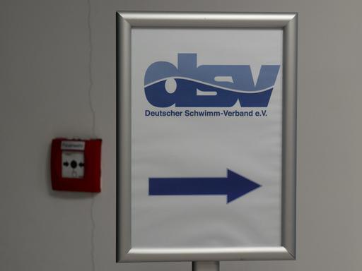 Auf dem Schild mit blauer Schrift auf weißem Grund zeigt ein pfeil nach rechts. Es hängt an einer Wand neben einem Feuer-Notmeldeknopf. 