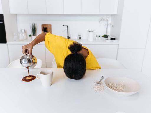 Eine erschöpfte Frau mit dem Kopf auf der Tischplatte verschüttet ihren Kaffee