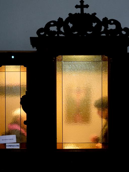 In einem Beichstuhl sitzen zwei Personen, die durch die getrübte Scheibe nicht zu erkennen sind.