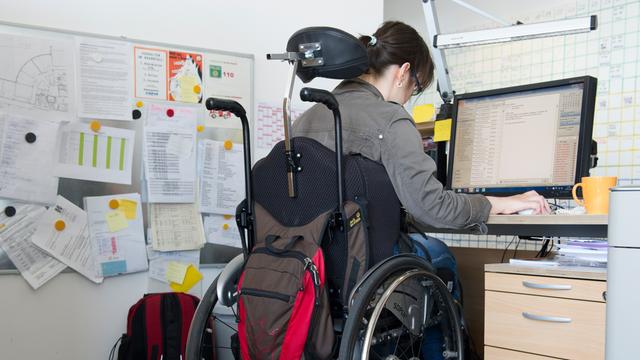 Eine Frau im Roll-Stuhl sitzt in einem Büro an einem Schreib-Tisch.