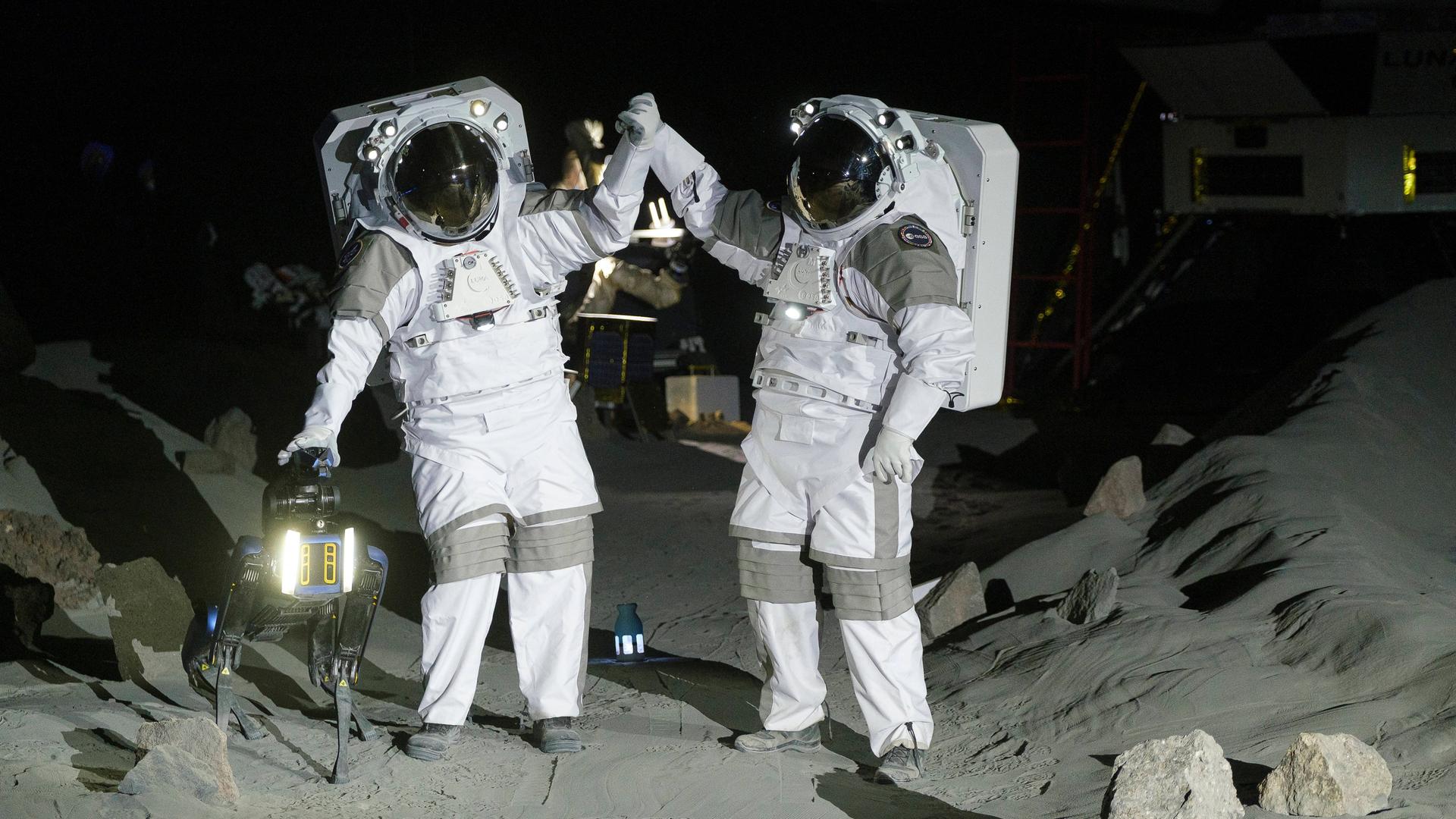 Zwei Astronauten stehen in Raum-Anzügen in einer Mond-Landschaft und schlagen mit den Händen ein.