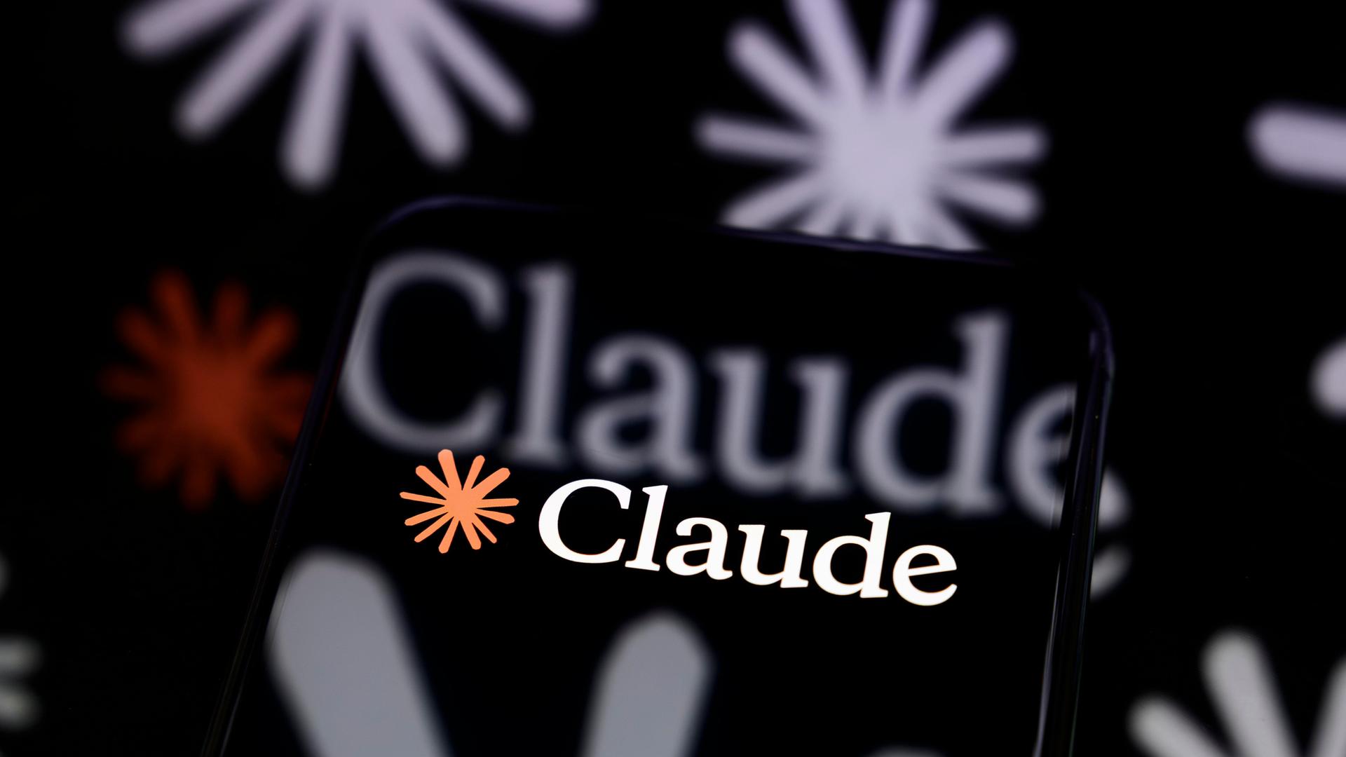 Das Claude-Logo ist auf dem Bildschirm eines Smartphones zu sehen, das auf einer reflektierenden Fläche steht 