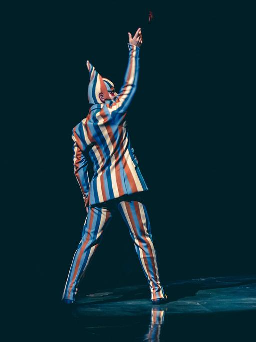 Ein Mann in einem gestreiften Clownsanzug steht gerade in einer Manege und hält den rechten Arm hoch. 